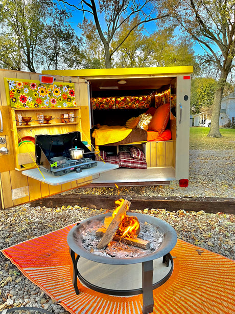 Relaxing RV rental setup: Open camper with cozy bed & outdoor kitchen. A lit fire pit sits on an orange mat.