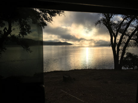 Stunning sunrise over a misty lake, seen from an RV rental window. Features silhouetted trees, a small boat, and bright sun reflection.