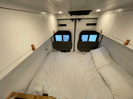 Bright RV rental interior: large white-covered bed, custom overhead cabinets with leather pulls, rear windows, and ceiling lights.