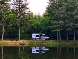 Serene rv rental camper van reflecting in a peaceful lake, surrounded by towering trees and a picnic area.