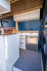 RV rental interior featuring a modern kitchen with white cabinets, fridge, and wood countertops, plus a brown leather bench and slatted wood ceiling.