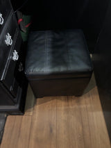 Black leather-look storage ottoman on a wood floor in a compact RV rental setting, beside a dark dresser.