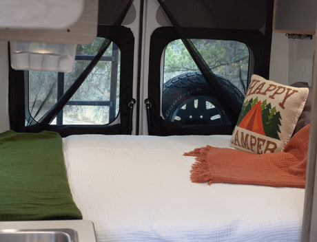 Cozy RV rental interior: bed, "Happy Camper" pillow, and scenic view through mesh windows.
