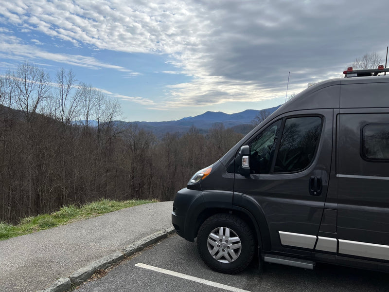  colorful sunset sky.|Dark grey Ram ProMaster RV rental parked curbside by a grand stone mansion with an arched entrance.|A grey RV rental camper van is parked — photo 9