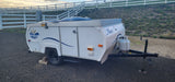 A white Aliner Sportliner pop-up RV camper parked on gravel with a fence and grassy hill behind. Perfect for your rv rental!
