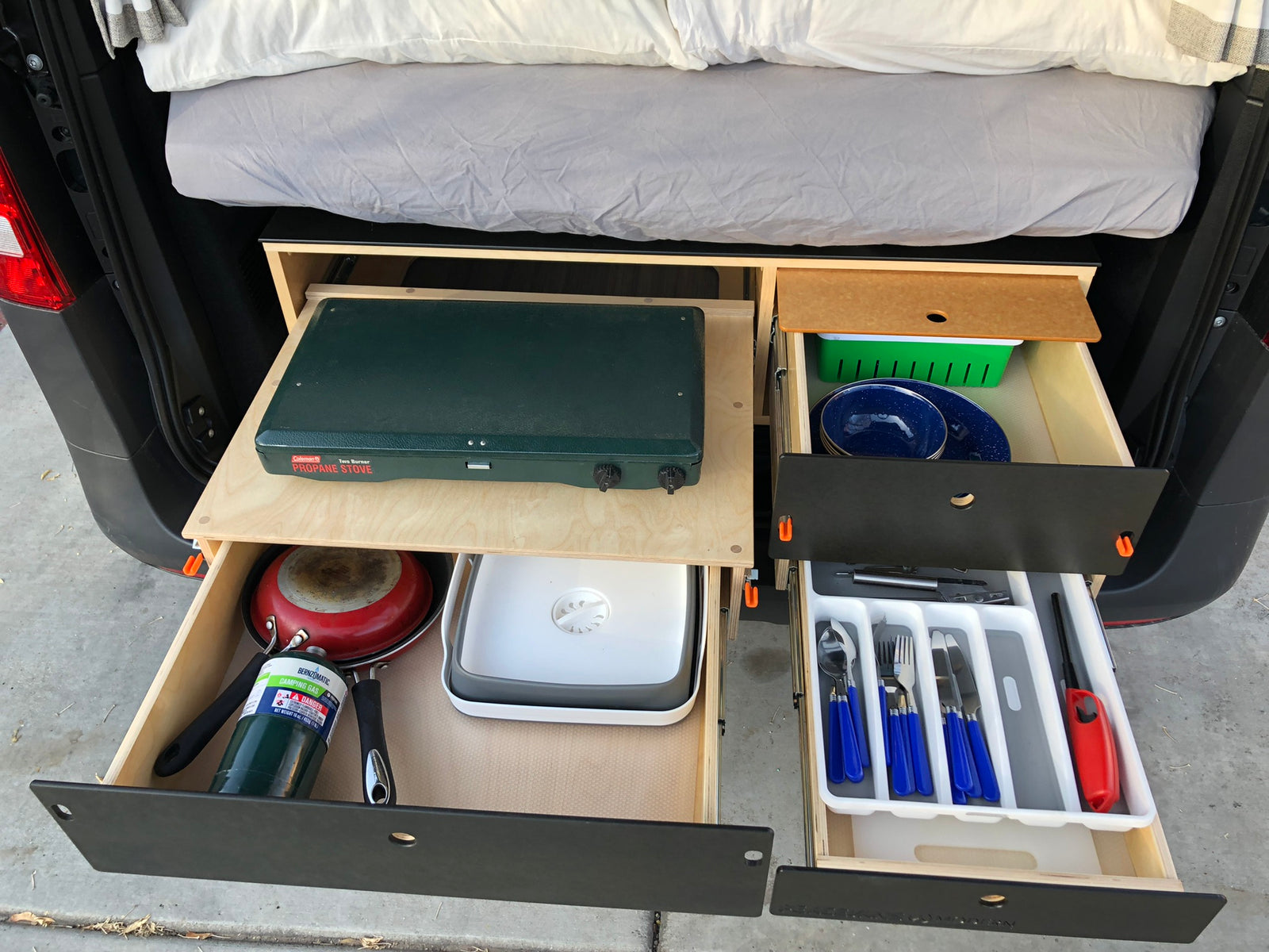 RV rental's pull-out kitchen: Coleman stove, pan, plates, and blue utensils in organized drawers.