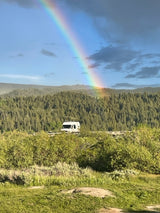 A vibrant rainbow arcs over a forest, with an RV nestled in the green foreground. Explore nature's beauty with an RV rental.