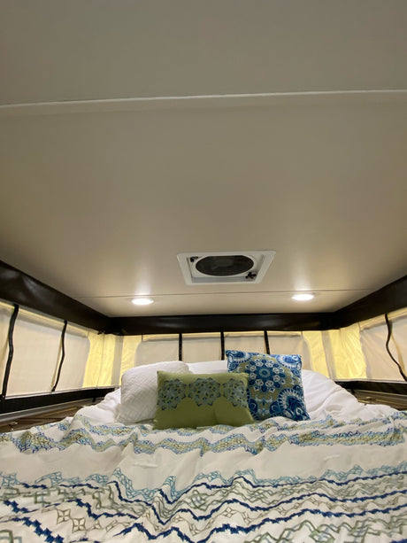 RV rental: Plush bed with patterned duvet, cozy pillows, ceiling fan, and bright lights in a pop-top camper interior.