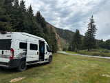 Serene nature spot! A white camper van rv rental sits by a clear mountain stream, surrounded by tall pine trees.