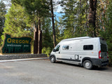 Gray camper van parked by the iconic Oregon welcomes sign in a forest setting, ideal for an RV rental road trip.