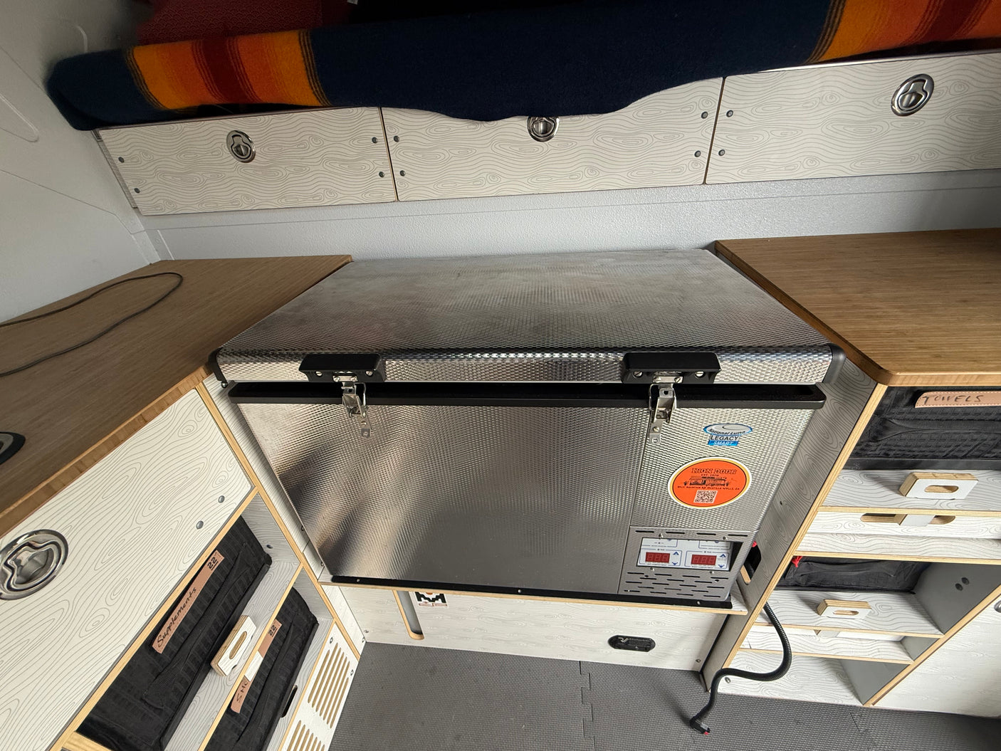 RV rental van interior showcases a portable fridge, sleek patterned storage cabinets, and an overhead sleeping area.