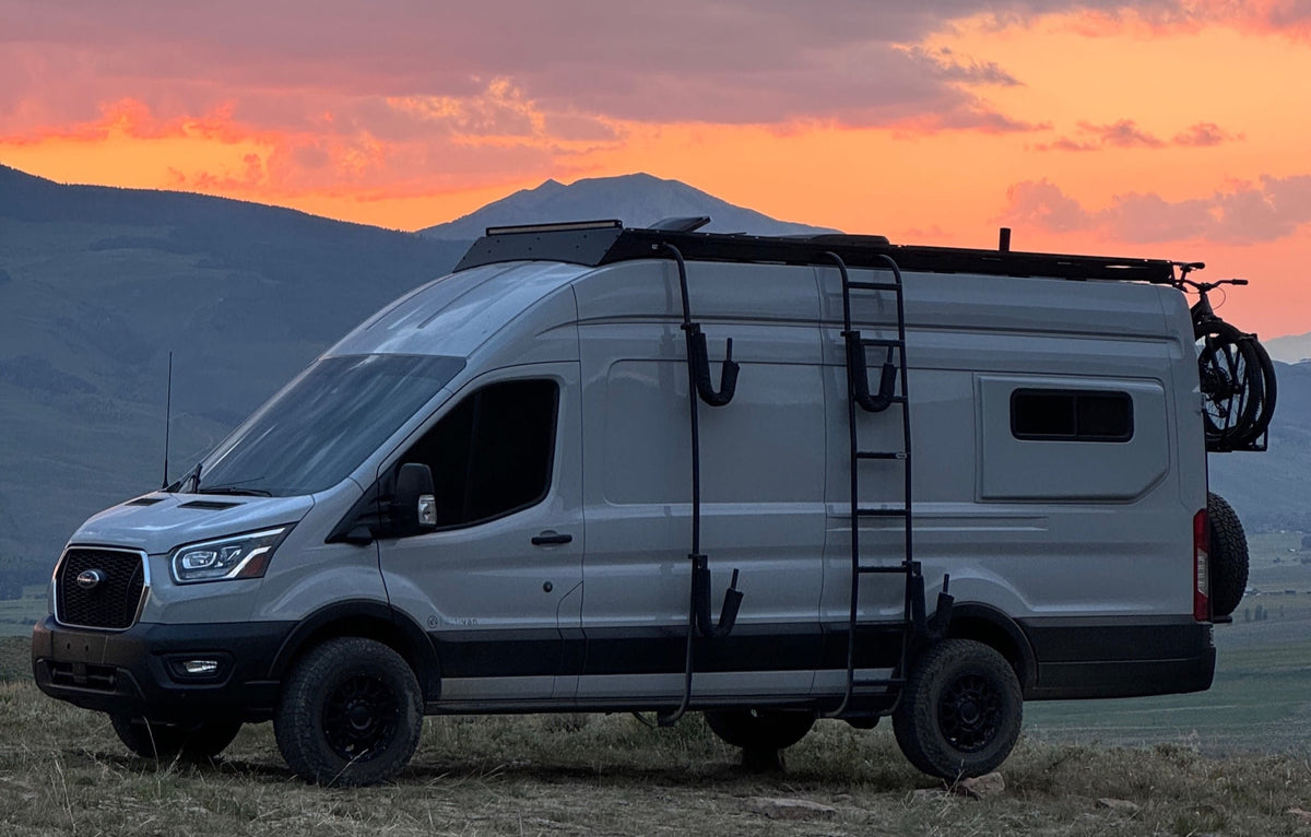 A gray Ford Transit camper van RV rental, equipped for adventure, is parked on a hill with mountains under a vibrant sunset.