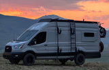 A gray Ford Transit camper van RV rental, equipped for adventure, is parked on a hill with mountains under a vibrant sunset.