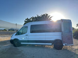 Bright sun on a white rv rental van with its rear door open, parked on sand against a mountain backdrop.