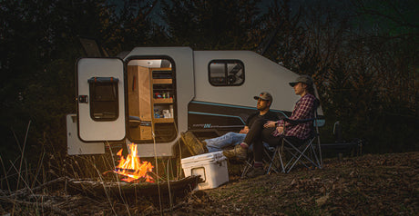 Two people relaxing by a cozy campfire next to a compact camper under a starry sky, perfect for an RV rental getaway.