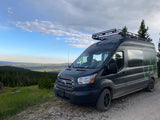 Topographic-wrapped RV rental van with a roof rack and light bar, parked on a dirt path with sweeping mountain views.