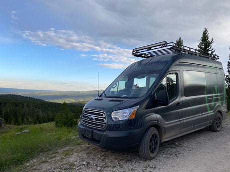 Topographic-wrapped RV rental van with a roof rack and light bar, parked on a dirt path with sweeping mountain views.