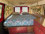 Inside this cozy RV rental: a comfortable bed with tropical print curtains and red cabinets, ideal for travel.