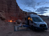 An RV rental camper van is parked amidst red rock formations at dusk, with a campfire and chairs set up for cozy desert camping.