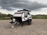 White Radical Bushwhacker RV rental parked on gravel, ideal for camping and outdoor adventures under a cloudy sky.