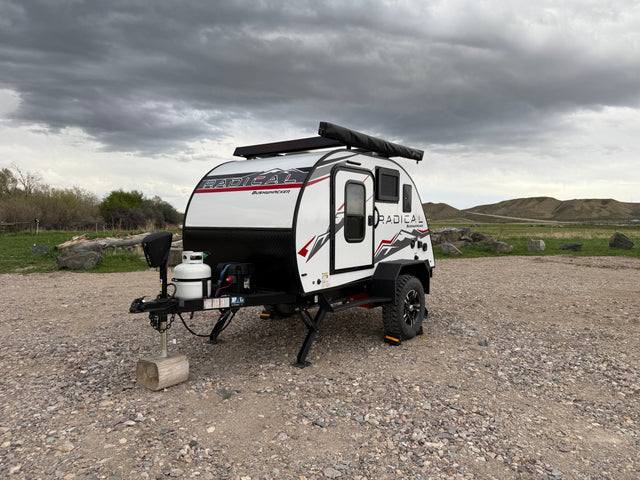 White Radical Bushwhacker RV rental parked on gravel, ideal for camping and outdoor adventures under a cloudy sky.