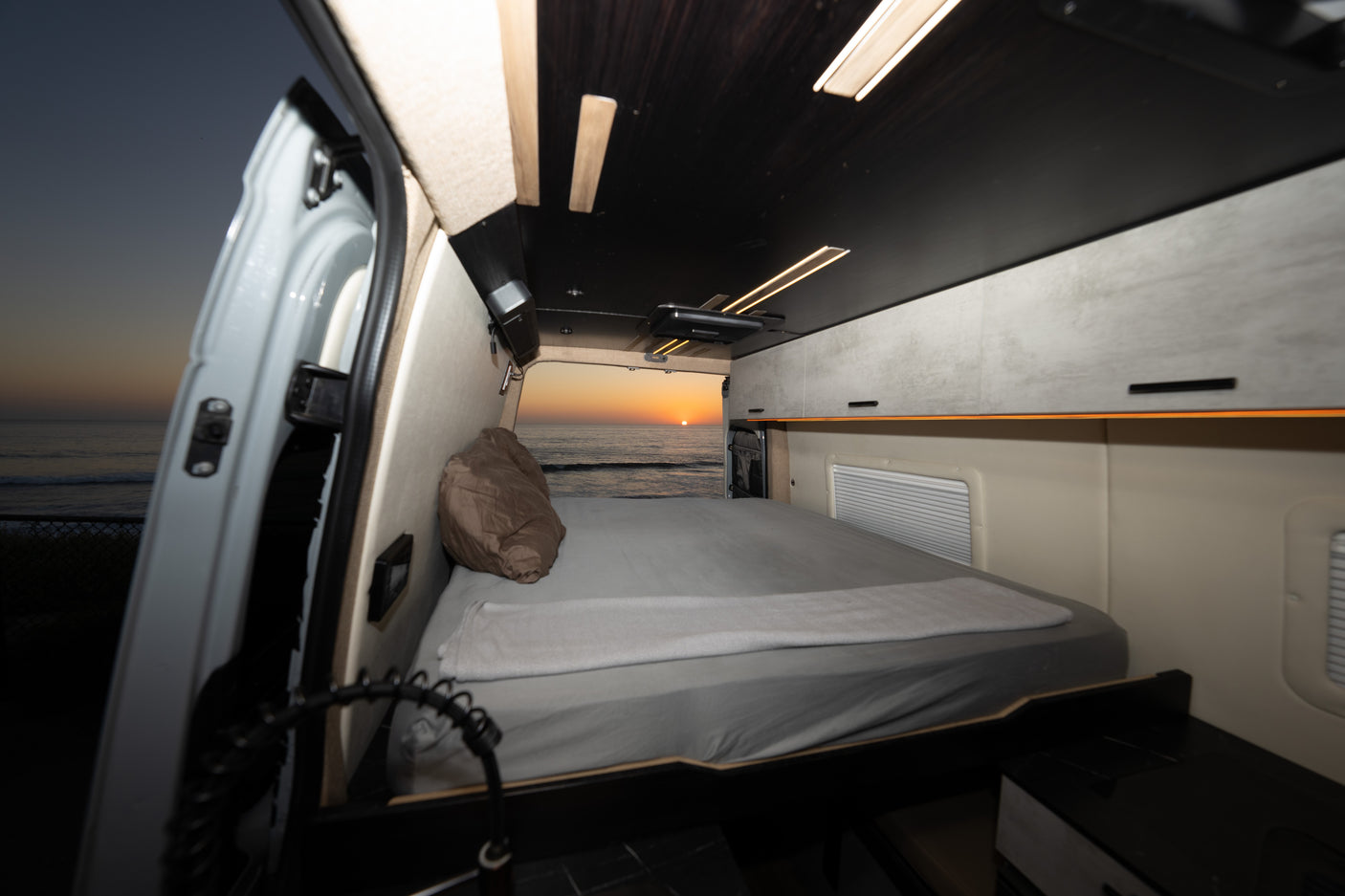 Experience an RV rental! Cozy camper van interior with bed facing a beautiful ocean sunset. Perfect for a relaxing van life escape.