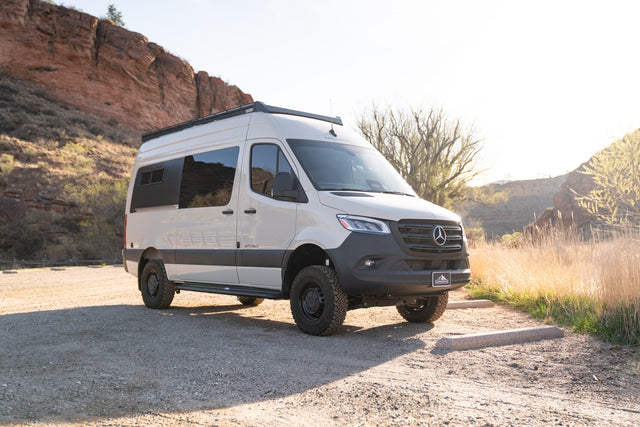 Mercedes-Benz Sprinter camper van for rv rental, parked on gravel with rocky cliffs and dry brush in a scenic off-road setting.