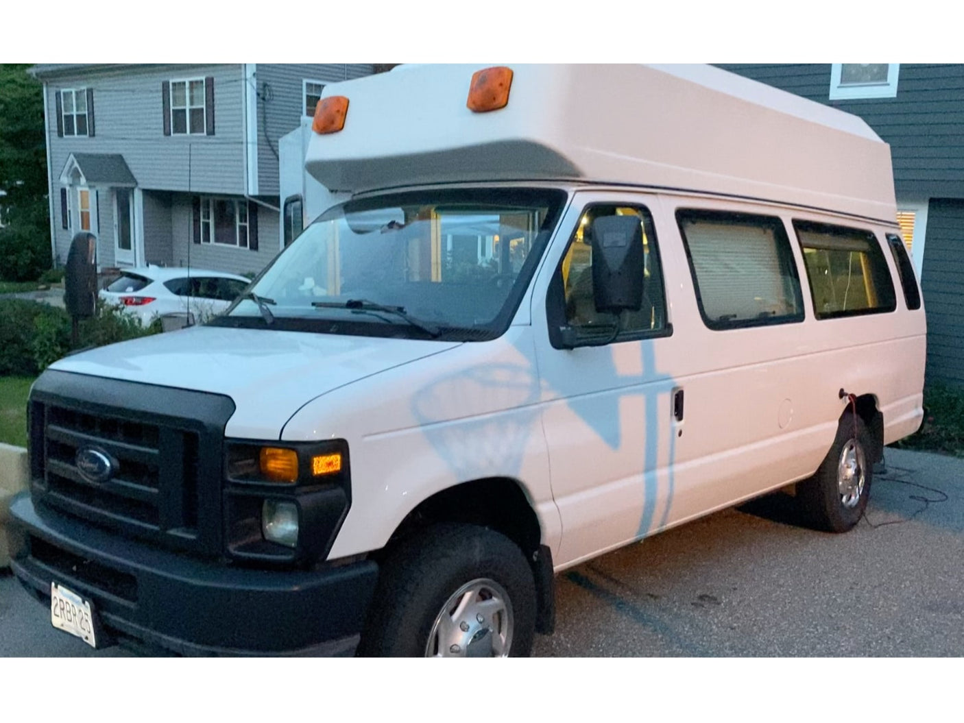 White Ford high-top van, resembling an RV rental, parked on an asphalt driveway in a residential area.