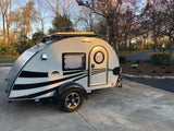 A stylish teardrop RV rental, grey with black stripes, parked on a driveway. Ready for camping adventures with solar power.