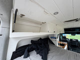 Cozy RV rental interior with bed & overhead storage. Front cab visible, perfect for travel.
