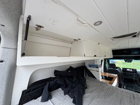 Cozy RV rental interior with bed & overhead storage. Front cab visible, perfect for travel.
