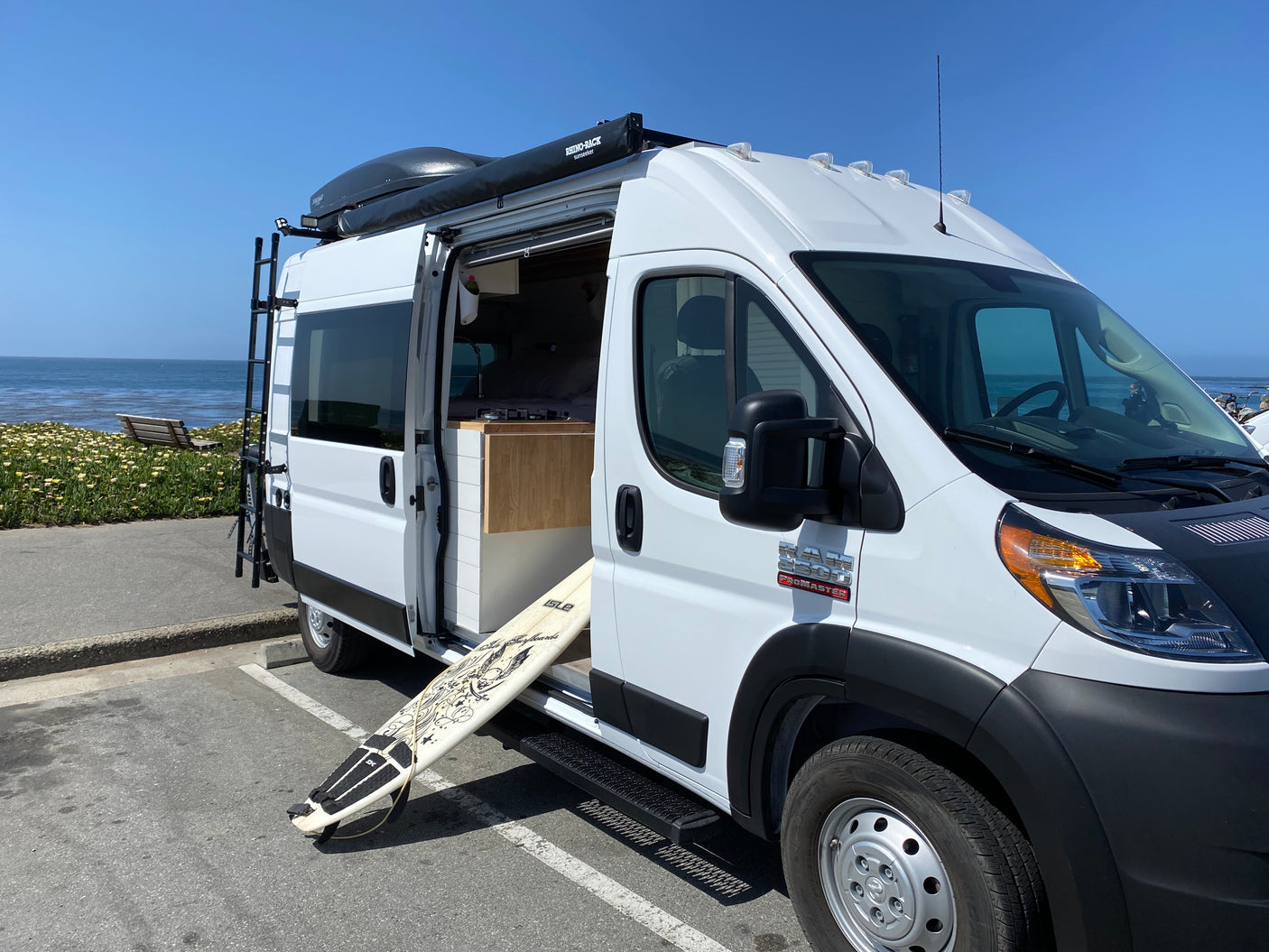 White Ram ProMaster camper van RV rental parked by the ocean with its side door open, revealing the interior and a surfboard.