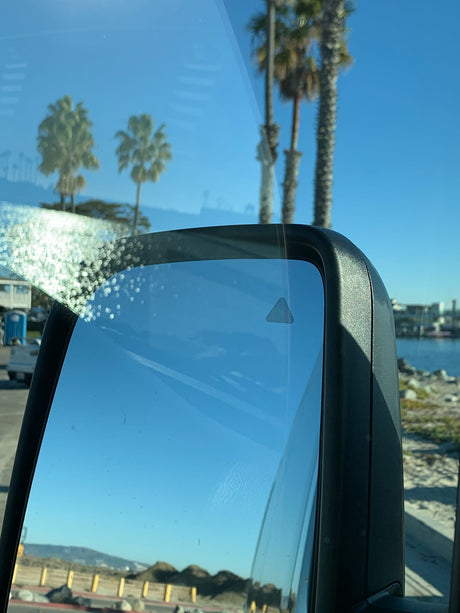 RV rental view: side mirror with blind spot monitor, palm trees & ocean under blue sky.