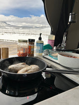 RV rental cooking in snow: Chicken sizzling on an induction cooktop inside, overlooking mountain scenery.