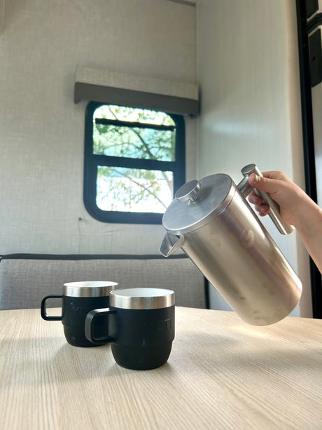Coffee time in an RV rental! A hand pours from a stainless steel French press into two black YETI mugs on a light wood table.
