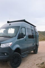 Adventure-ready blue camper van for rv rental, featuring rugged tires & roof access ladder, on a dirt road.