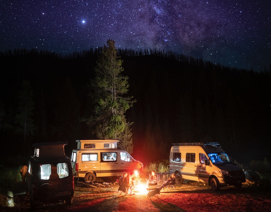 Night sky adventure with RV rental: Three camper vans, people by a campfire in a dark forest under brilliant stars.