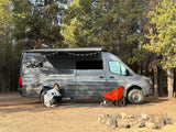 Cozy camping scene with an rv rental camper van. A person is wrapped in a blanket under the awning strung with lights at a campsite.