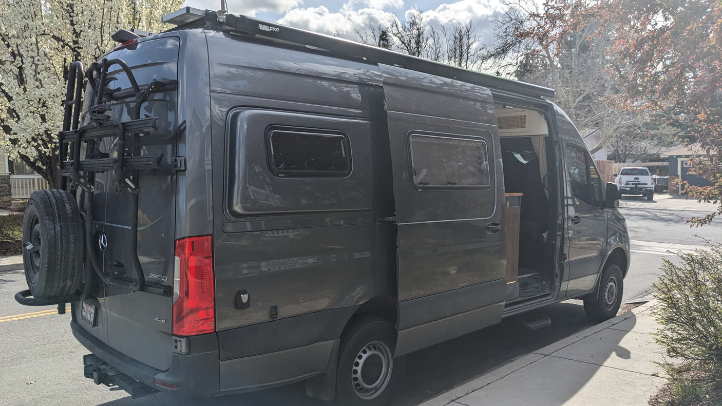 Gray Mercedes Sprinter camper van for RV rental, with bike rack, awning, and solar panels. Side door open.