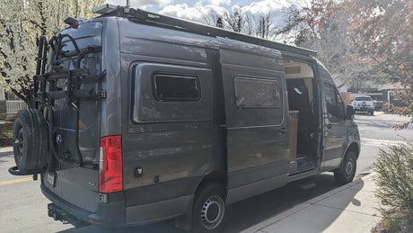 Gray Mercedes Sprinter camper van for RV rental, with bike rack, awning, and solar panels. Side door open.