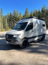 This white Mercedes-Benz Sprinter camper van, ideal for rv rental adventures, is parked in a sunny, forested setting.