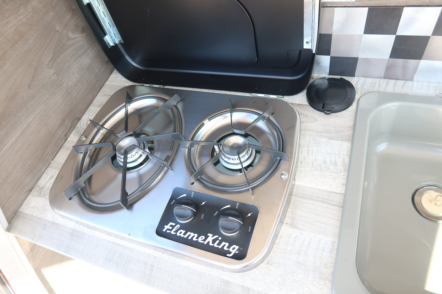 RV rental kitchen with stainless steel FlameKing two-burner stovetop and light gray sink, with an open lid.