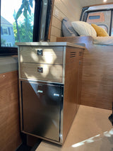 Inside a cozy rv rental: A camper van kitchen with an Isotherm fridge, drawers, and storage cabinet, next to a comfortable bed.
