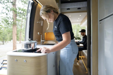 A woman cooks in an RV rental kitchen on an induction stove. A man works in the background by a forest view.