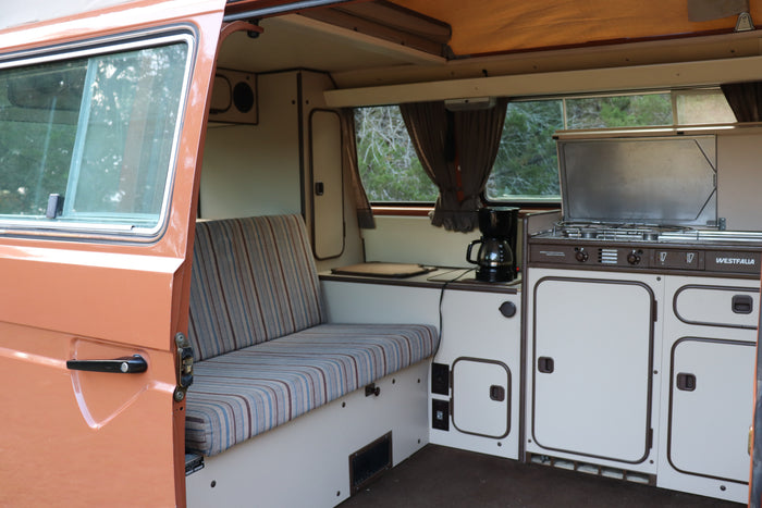Inside a retro Westfalia camper van, great for an RV rental. Features a striped bench, compact kitchen with stove, and storage.