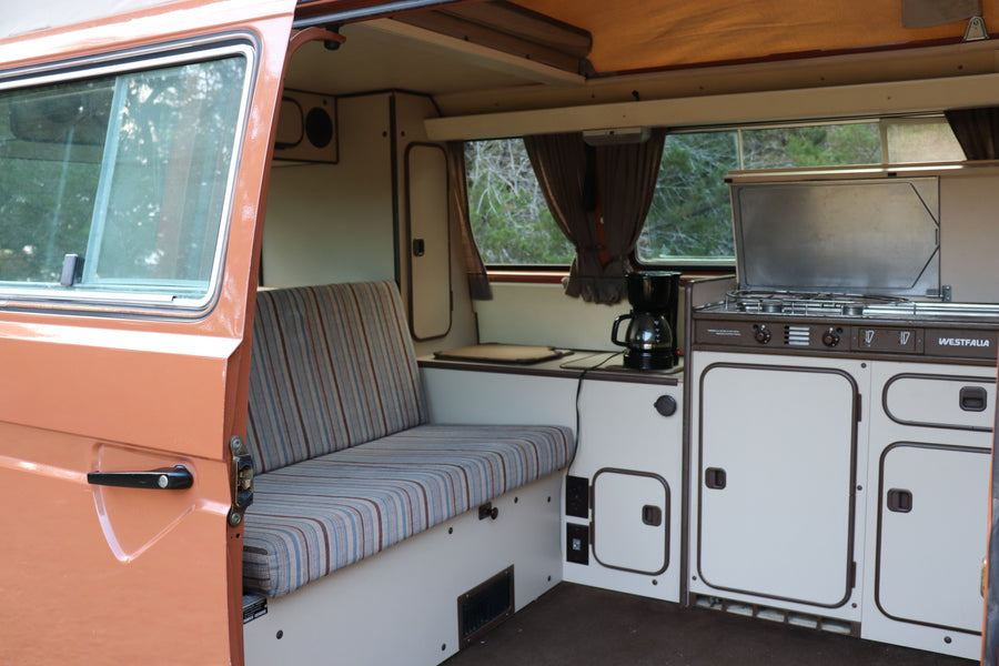 Inside a retro Westfalia camper van, great for an RV rental. Features a striped bench, compact kitchen with stove, and storage.