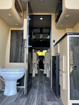 Modern RV rental interior with a toilet on the left, a kitchenette on the right, and passenger seating beyond.