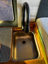 RV rental interior: Stainless steel sink, black faucet, ribbed drying rack, and wooden counter. Window and mattress visible.