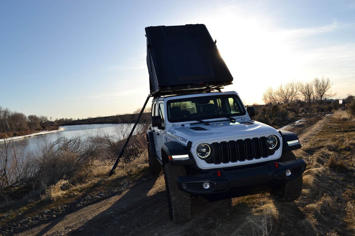 White Jeep Rubicon with a deployed rooftop tent, parked by a river at sunset. Ideal for an adventurous rv rental experience.