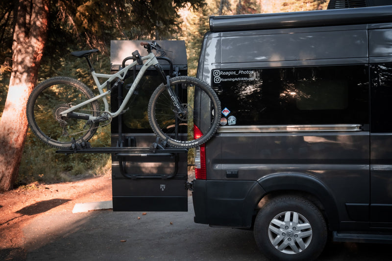 A dark grey RV rental, equipped with a Fezzari mountain bike on a Thule rear rack, is parked amidst a forest setting. — photo 4